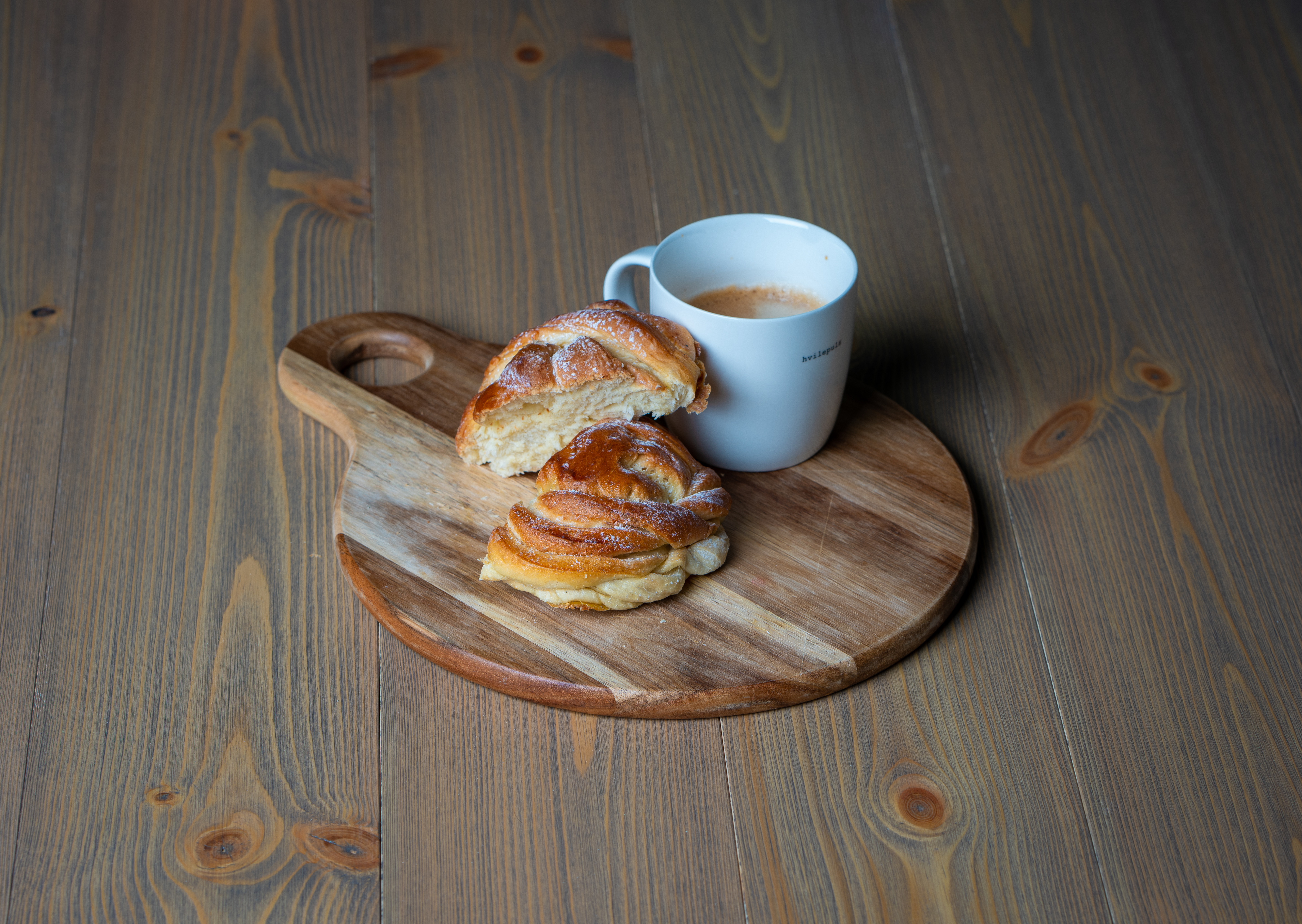 Et trebrett på gulvet med kanelboller og en kopp med kaffe