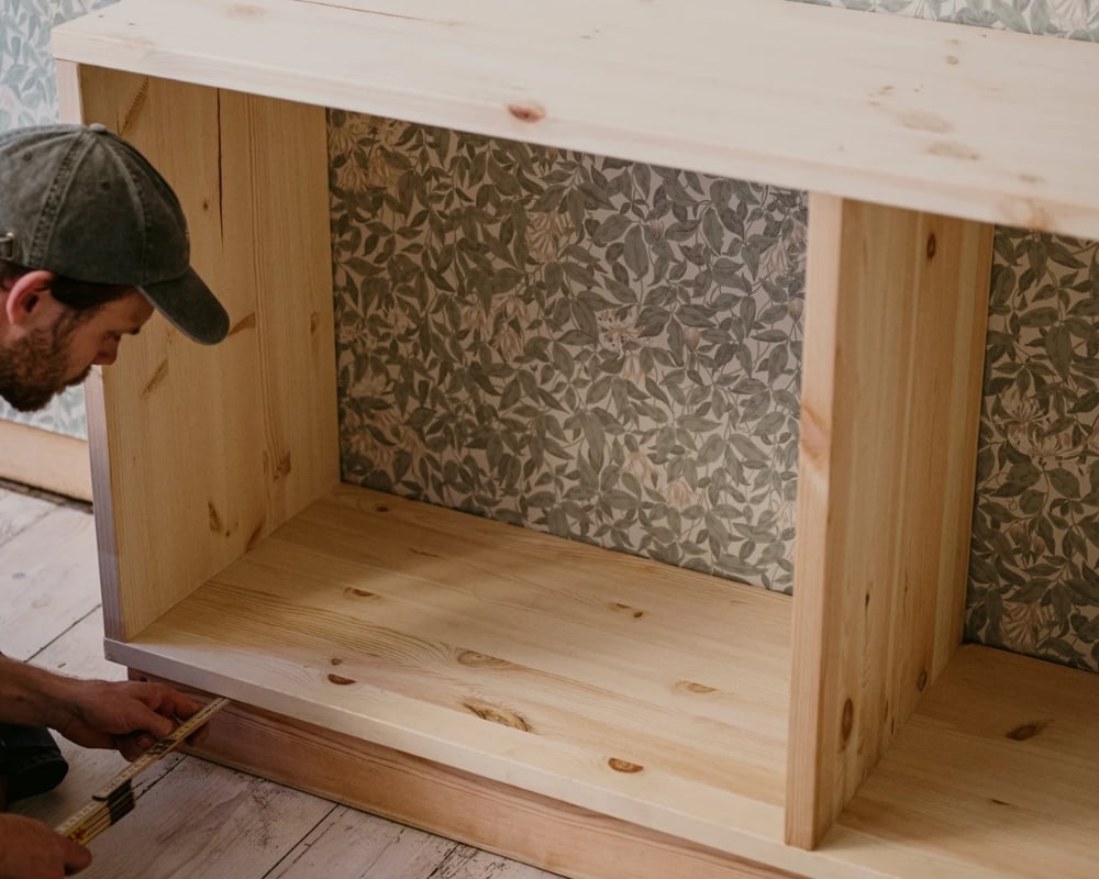 En man mäter avståndet från en ram till en hylla av trä, för att veta vart den ska skruvas fast