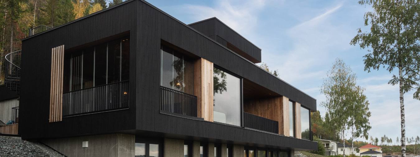 Moderne enebolig med mørk, nesten svart kledning i stående panel, kombinert med naturlige treelementer rundt store panoramavinduer. Huset er bygget på en betongbase med søyler og har balkonger med sorte rekkverk. I bakgrunnen ses løvskog i høstfarger og en blå himmel med lettskyet vær.