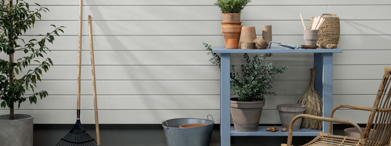 Hvitmalt, trekledning med liggende profil. Hagebenk, planter og hageredskaper plassert foran veggen