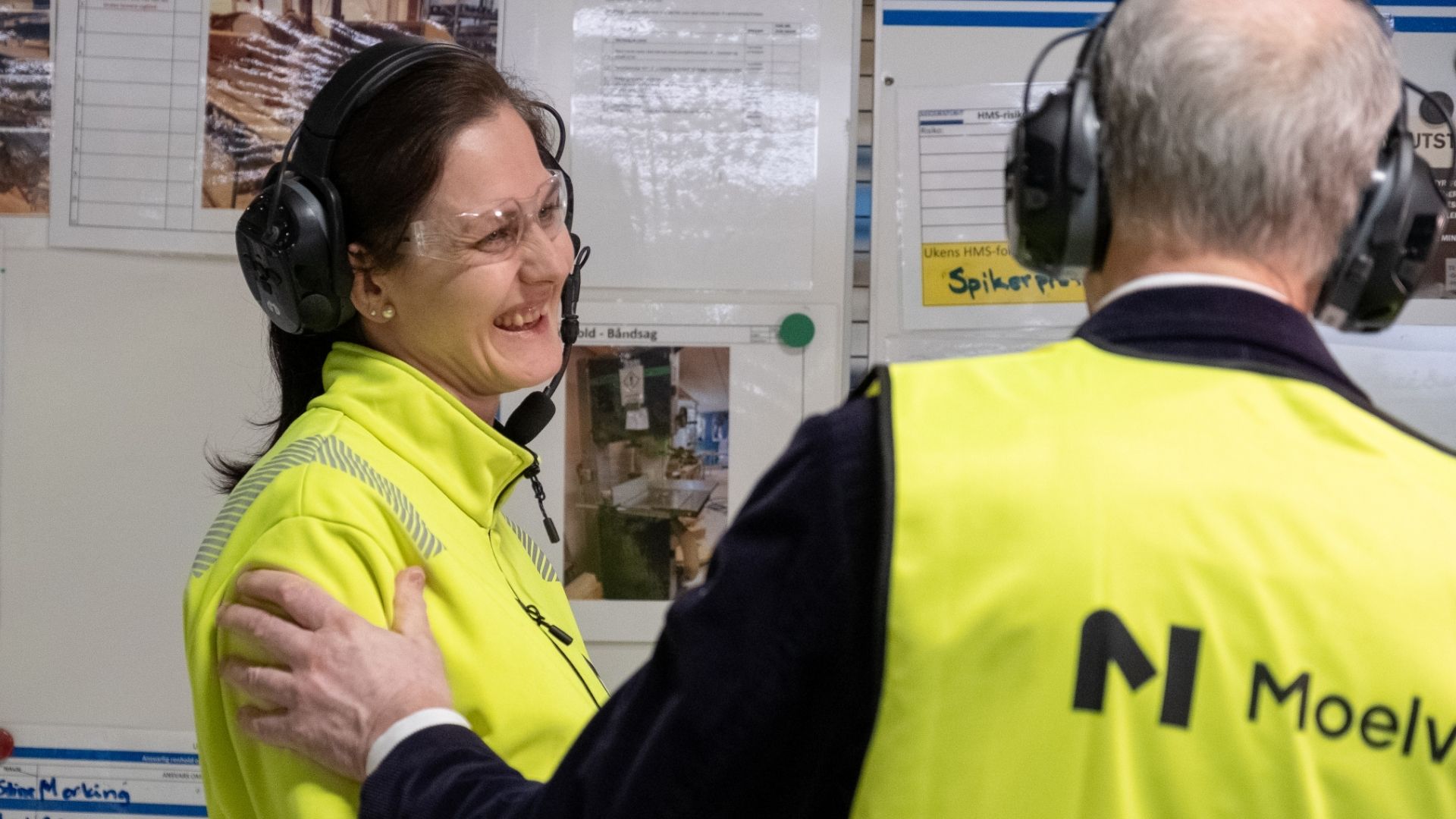 Kvinne i synlighetstøy smiler. Mann i synlighetstøy står med ryggen til og hoilder henne på armen.