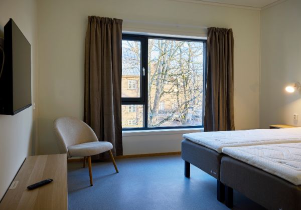 Rom med perspektiv mot en dobbelstseng, TV-benk og TV. Stort vindu der med gardiner.
