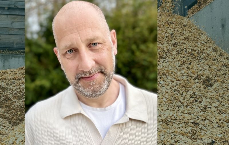 Närbild av av Carl-Gustaf Carlgren, stående utomhus med grönskande bakgrund. På båda sidor syns högar av träspån eller flis.