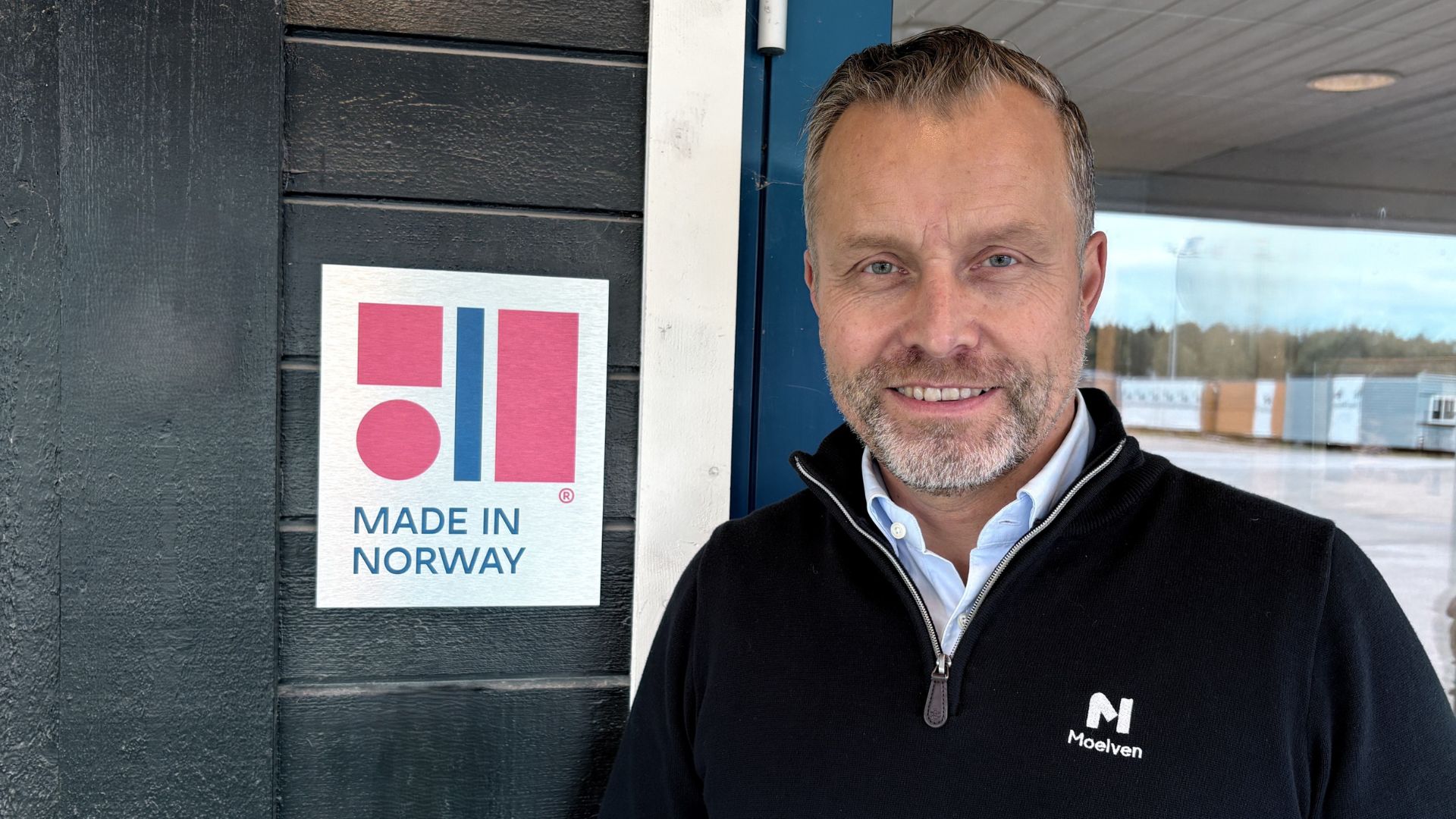 Smilende mann i Moelven-profilert genser står ved siden av et Made in Norway-skilt