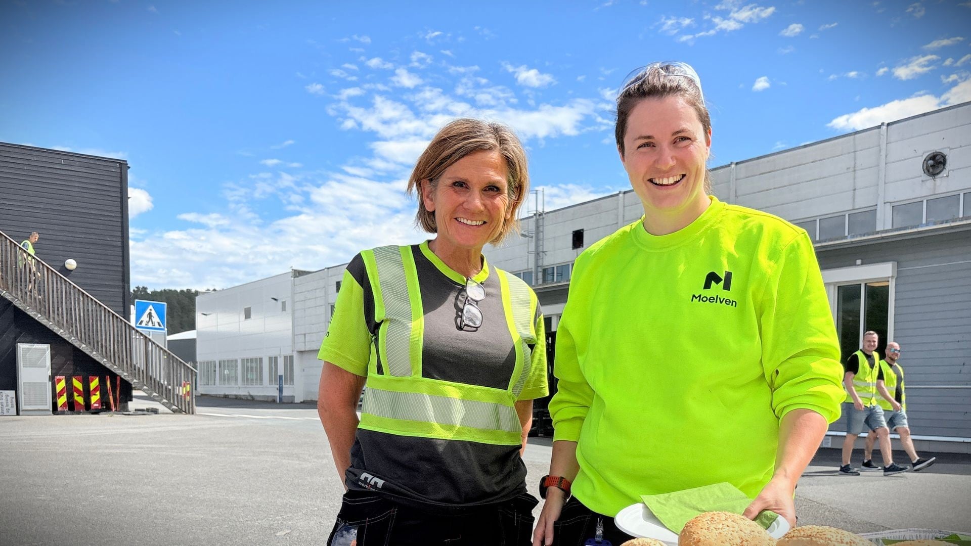 To smilende kvinner i synlighetstøy utenfor Mo9elven Byggmodul sine fabrikklokaler på Moelv.