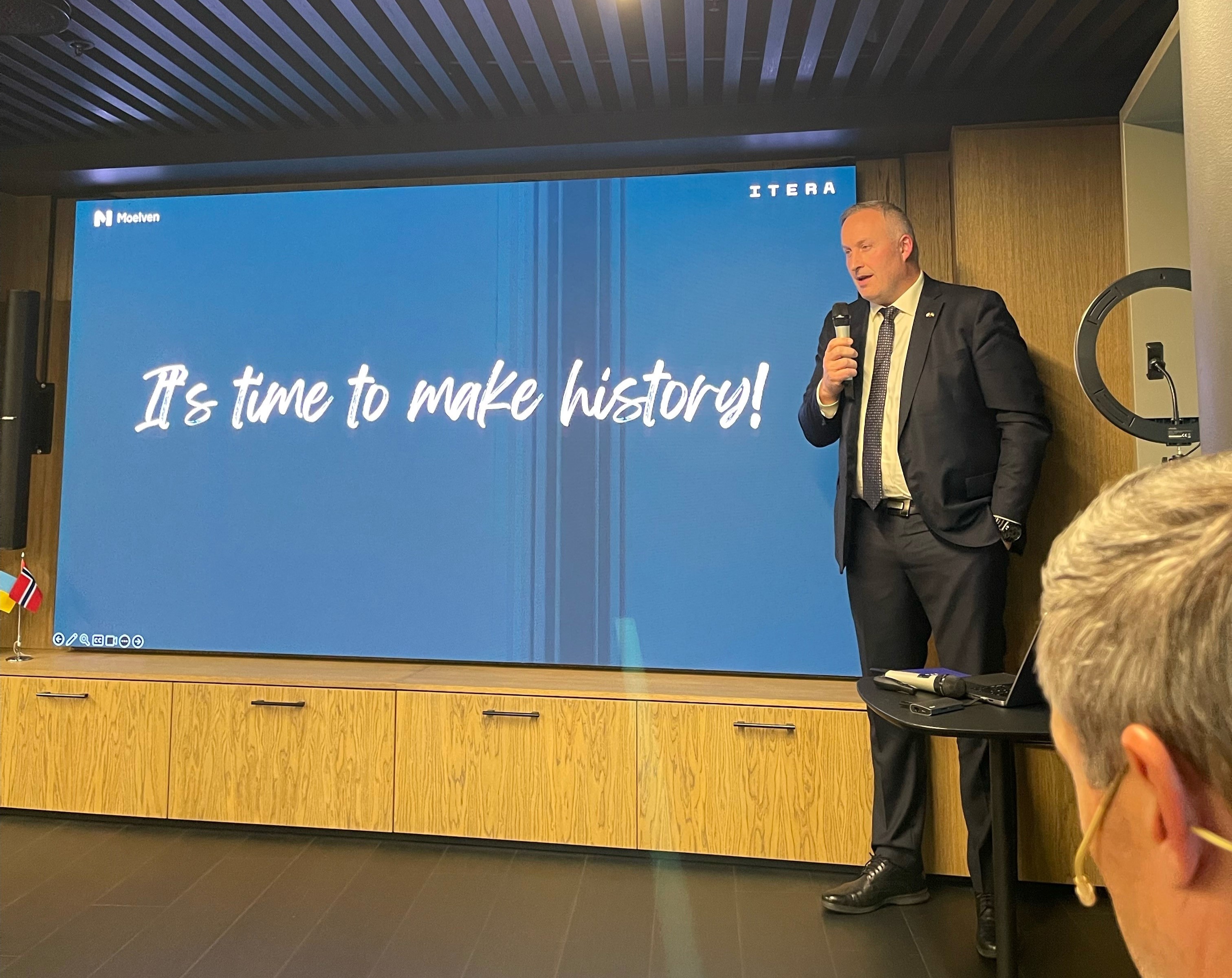 Dresskledd mann står ved en stor skjerm og snakker til en forsamling. Han holder en mikrofon i hånden.