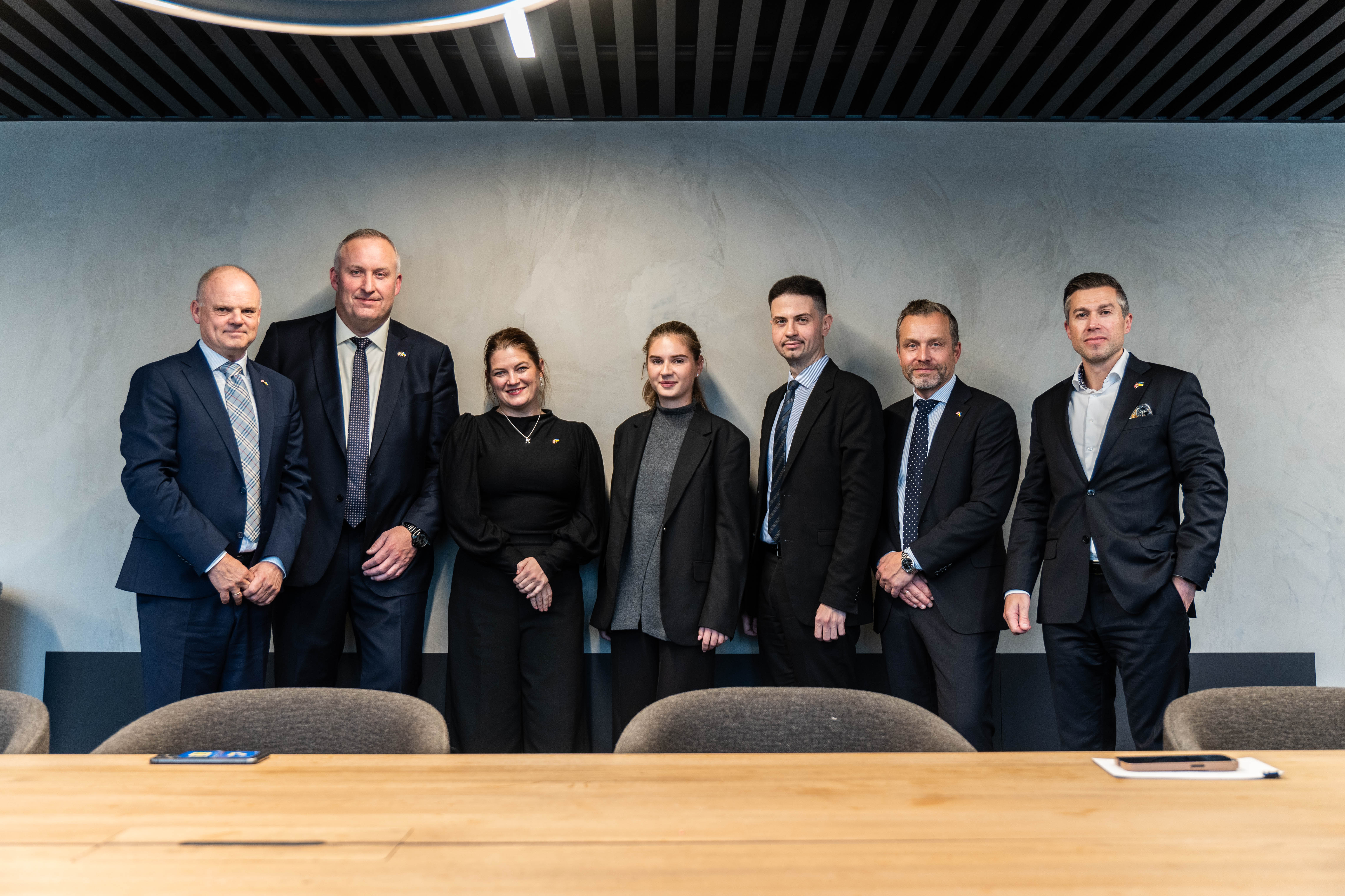 Gruppebilde av syv smilende og pyntede personer som står inntil en vegg.