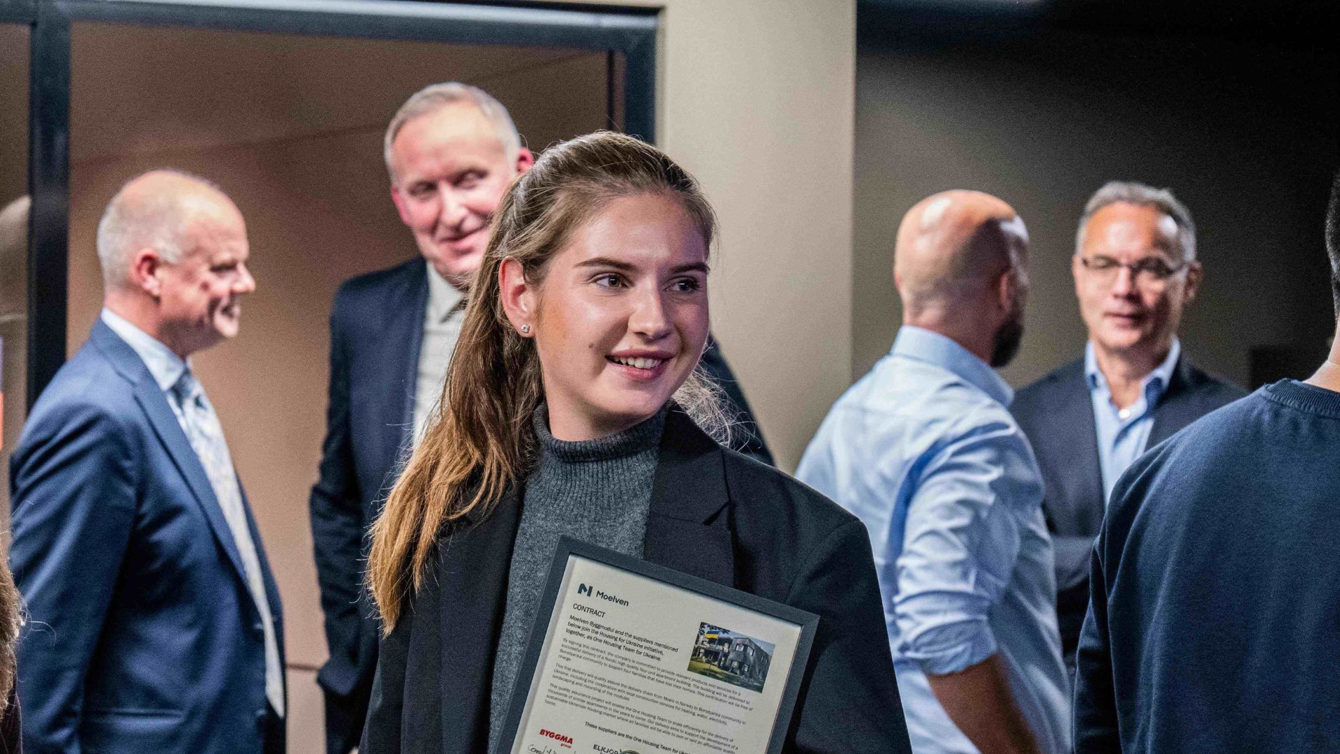 Smilende, ung kvinne med en signert, innrammet kontrakt i hånden. Hun er omgitt av blide mennesker.
