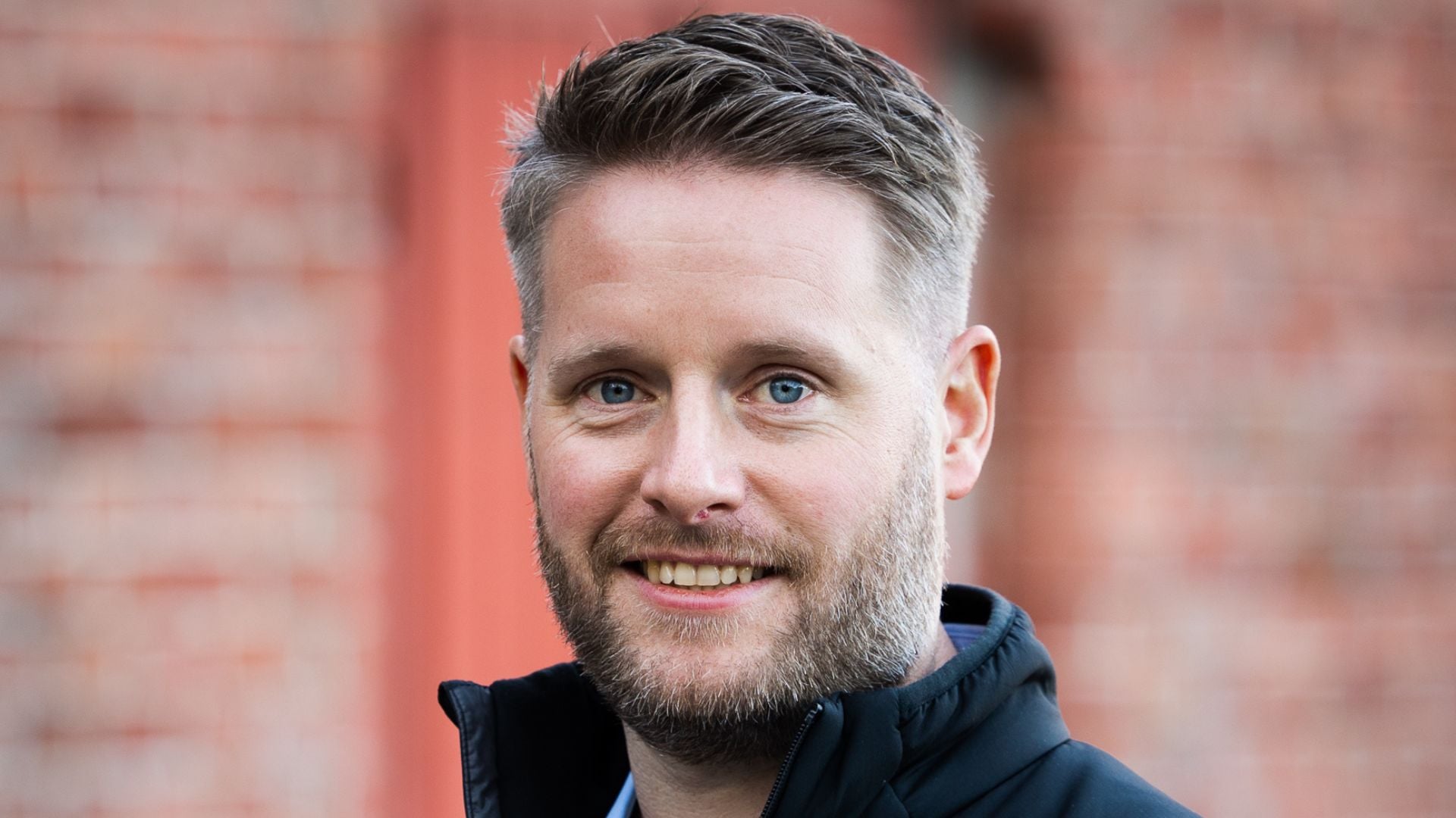 Smilende mann som ser i kamera. Teglstegnvegg i bakgrunnen.