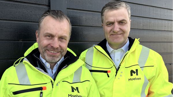 To smilende menn i Moelven synlighetsjakker står inntil en brun vegg.