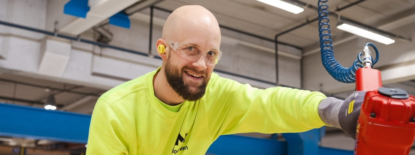 Smilende mann i fabrikk ser i kameraet og holder en spikerpistol.