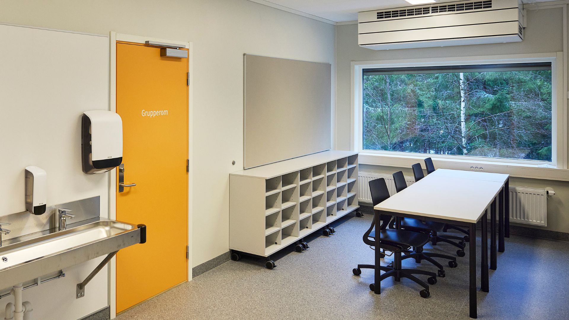 Klasserom som viser pulter, hyller, vask og en gul dør.