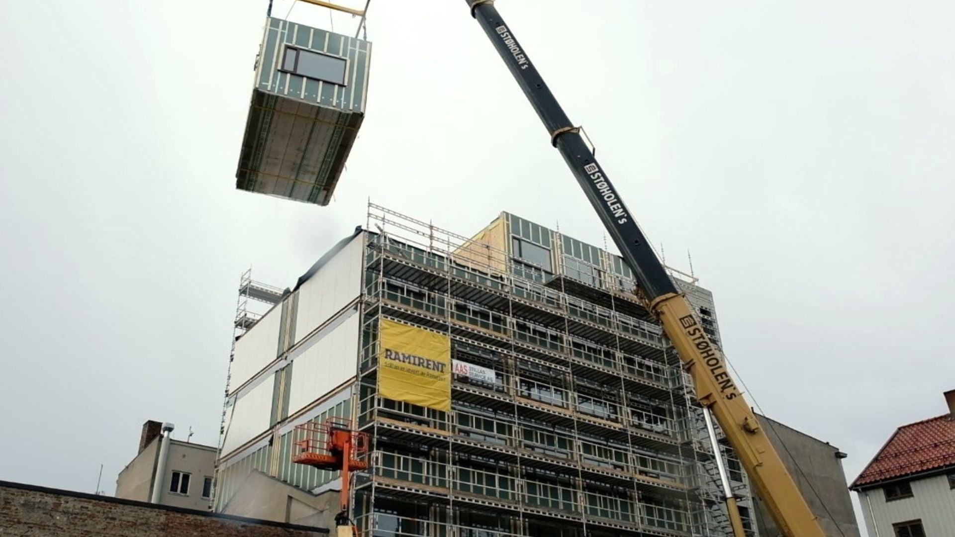 En byggmodul som henger i ei kran og skal løftes på plass på et modulbygg.