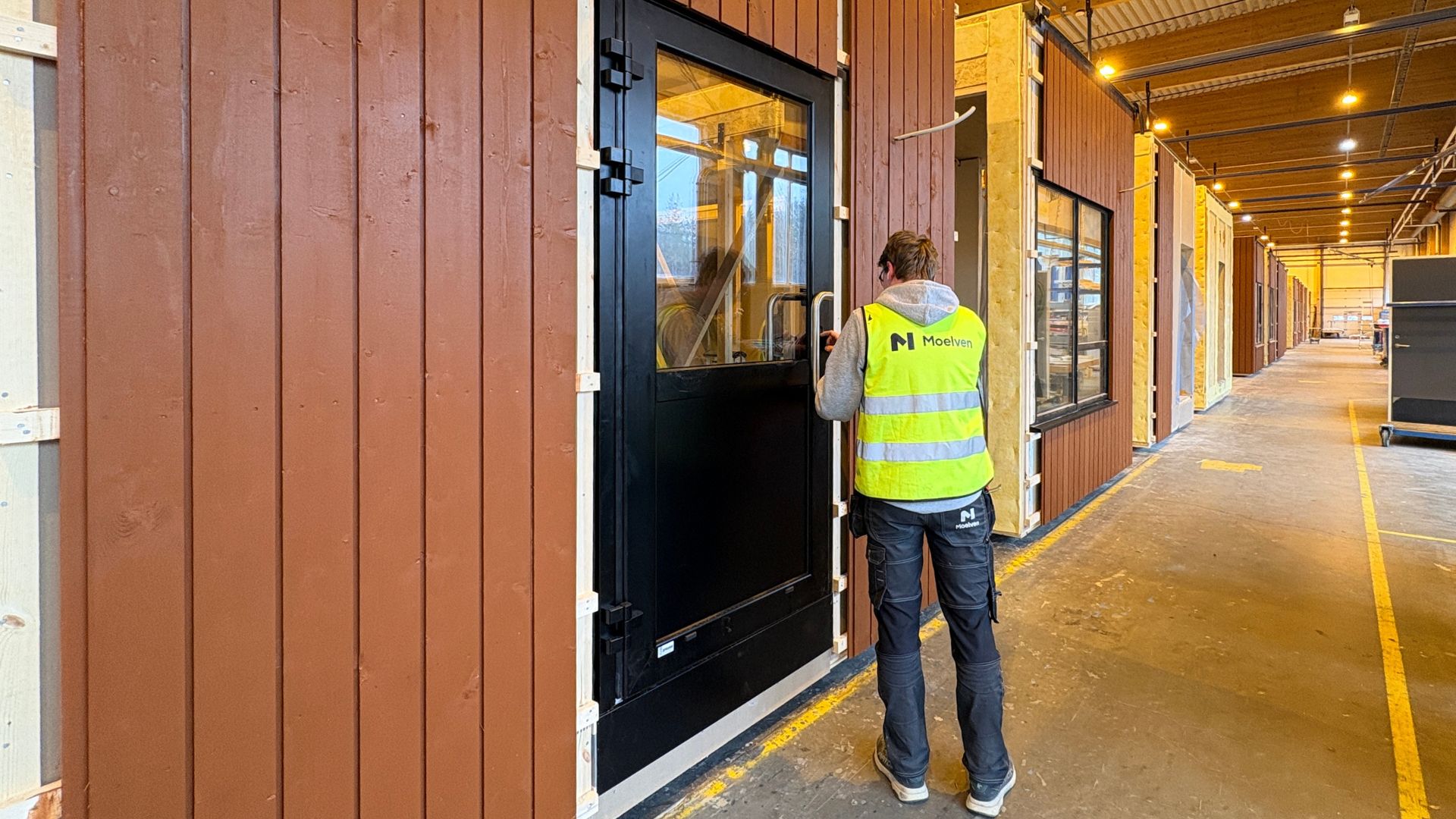 Medarbejder i refleksvest inspicerer et præfabrikeret træmodul i produktionshallen hos Moelven Byggmodul. Modulet har træbeklædt facade, sorte vinduer og dør og står på række sammen med andre moduler under produktion.