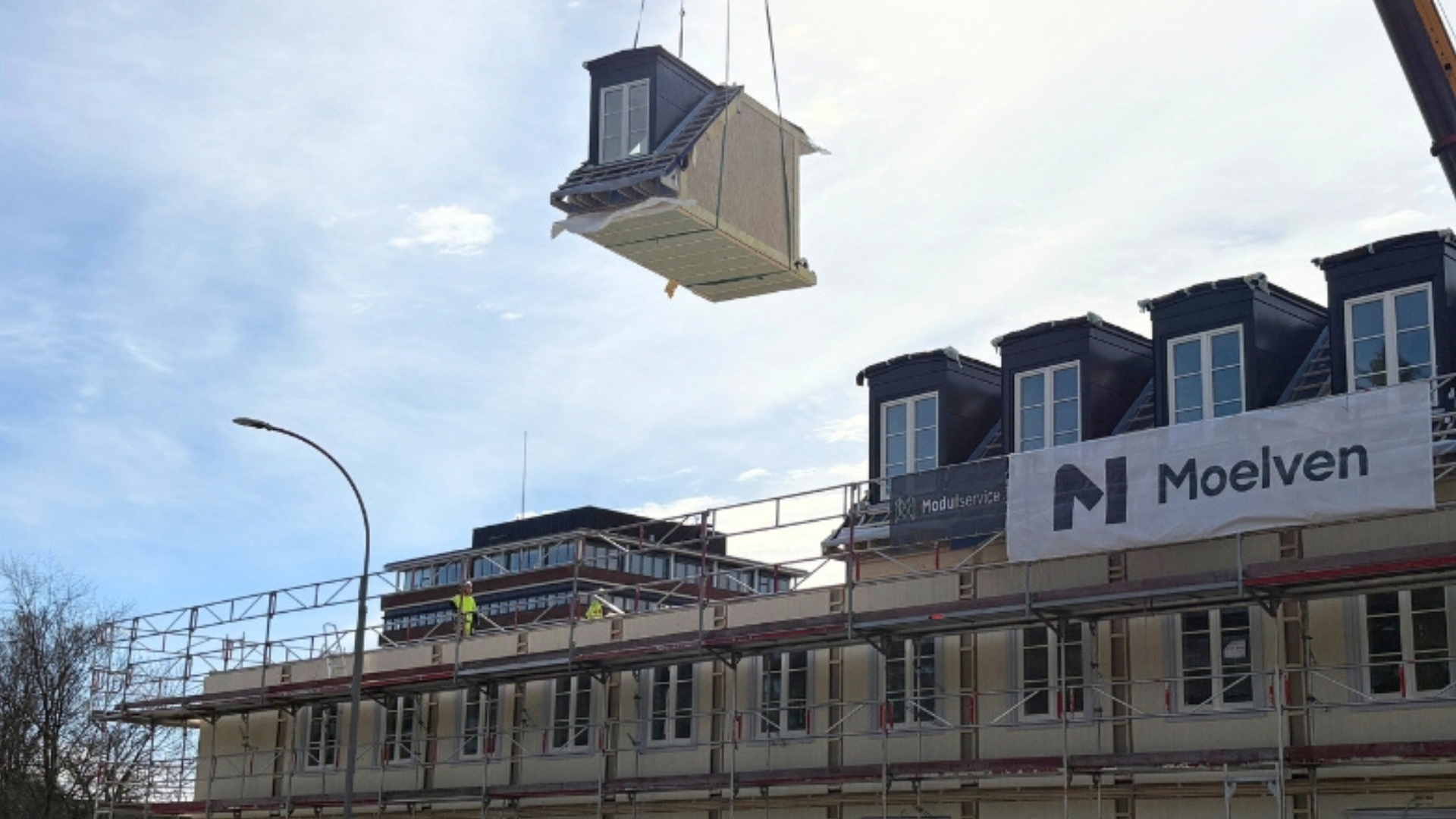 A wooden module suspended in the air by a crane above a modular building site.