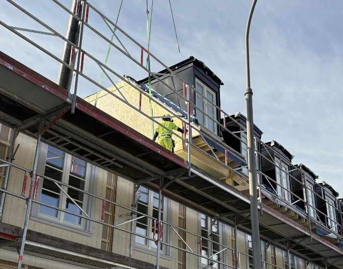 En byggmodul som heises på plass oppå et modulbygg med stillas. Arbeider med synlighetstøy og hjelm står på bygget.
