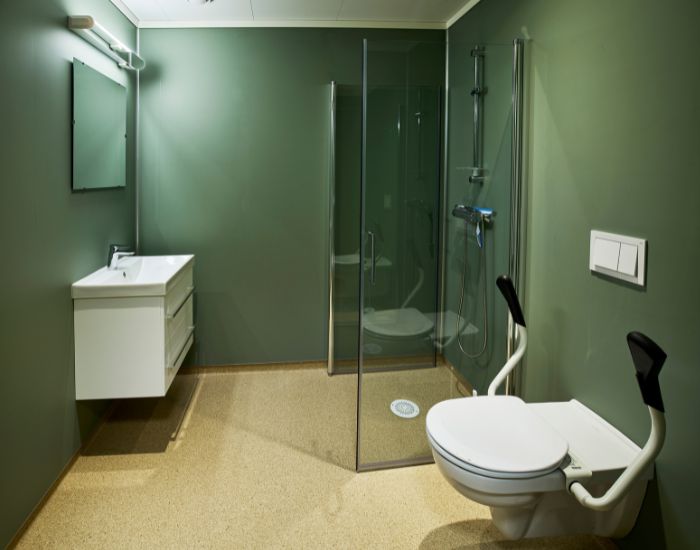 Fra baderom med handicap-toalett i front. Grønne vegger, okergule gulv, dusjvegger og hvite baderomsmøbler.