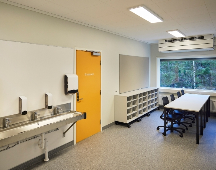 Klasserom med vask, reol og pulter. Gul dør inn til annet rom.