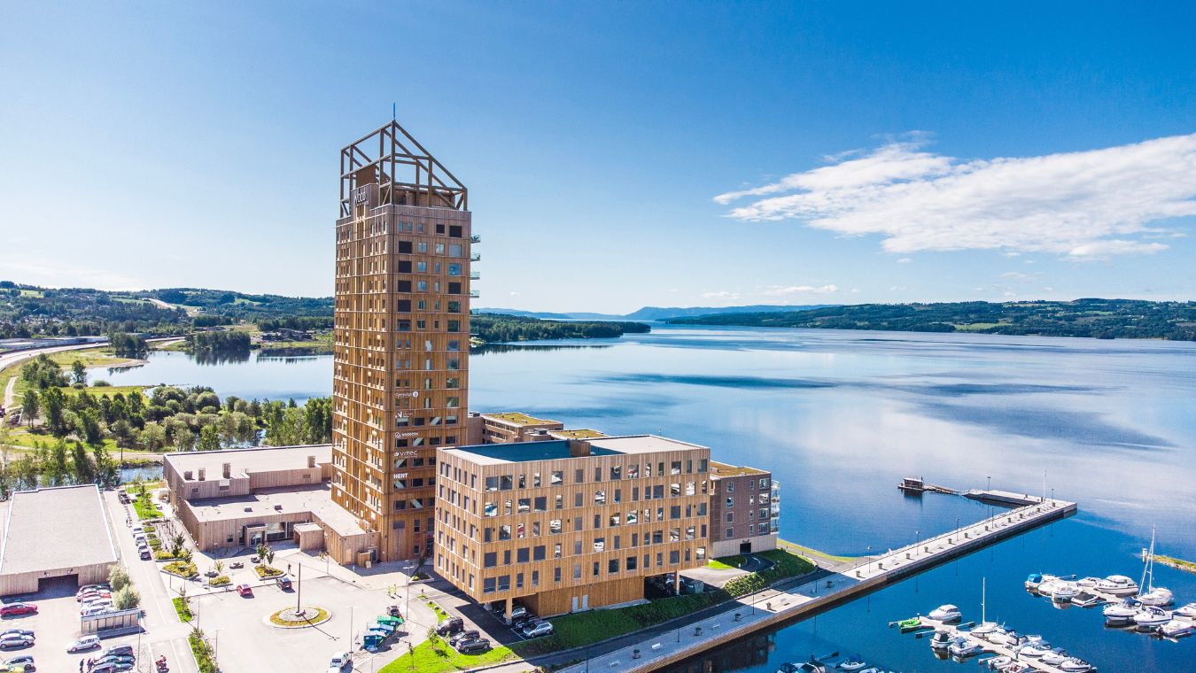 Mjöstårnet med övriga byggnader vid mjöskanten fotad i sol och blå himmel.
