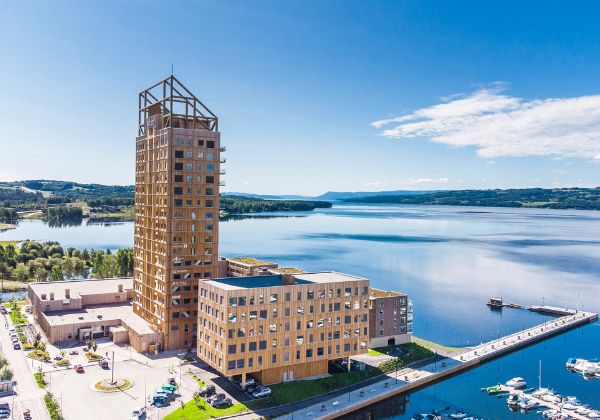 Mjöstårnet med övriga byggnader vid mjöskanten fotad i sol och blå himmel.