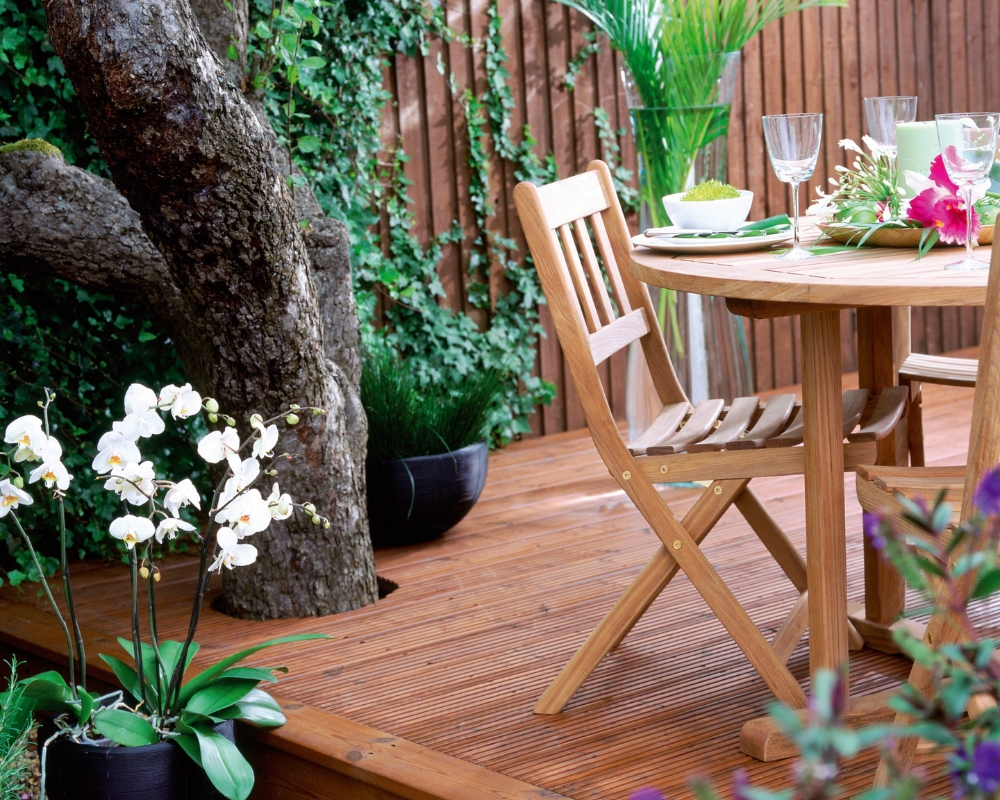 En trall av ThermoWood, flertal växter och ett träd, samt stolar och bord med dukning
