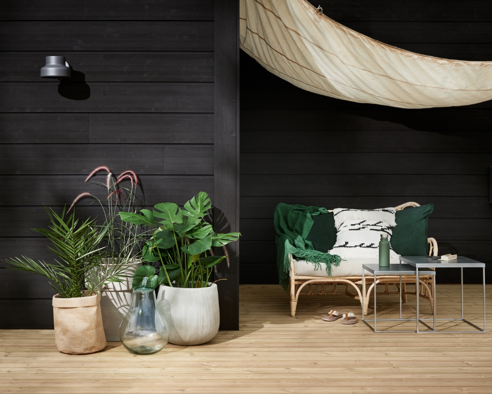 En svart limträpanel och trall, samt utomhusmöbler i form av soffa, bord, hängmatta och växter