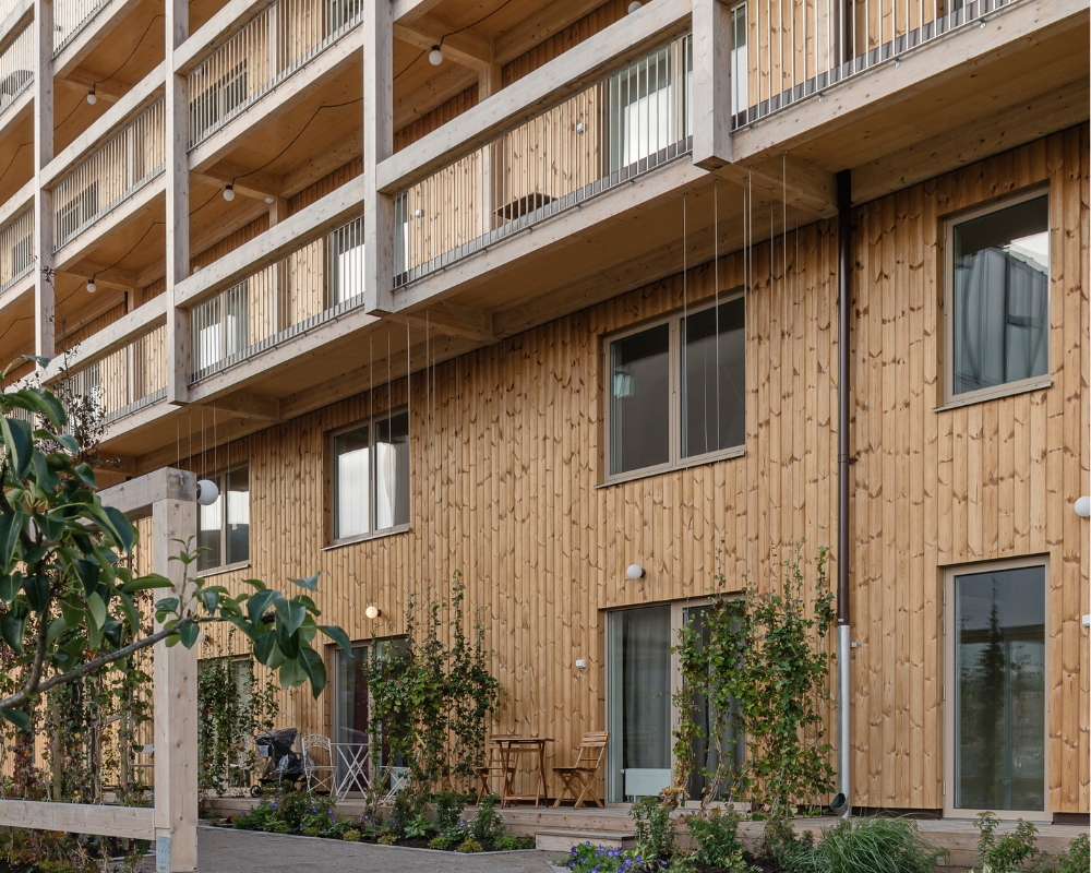 Ett lägenhetshus med träpanel, samt växter framför huset