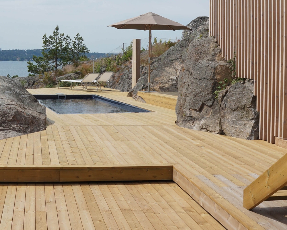 En altan med trall byggd runt stenar, en pool och solsängar & parasoll, samt utsikt mot vatten