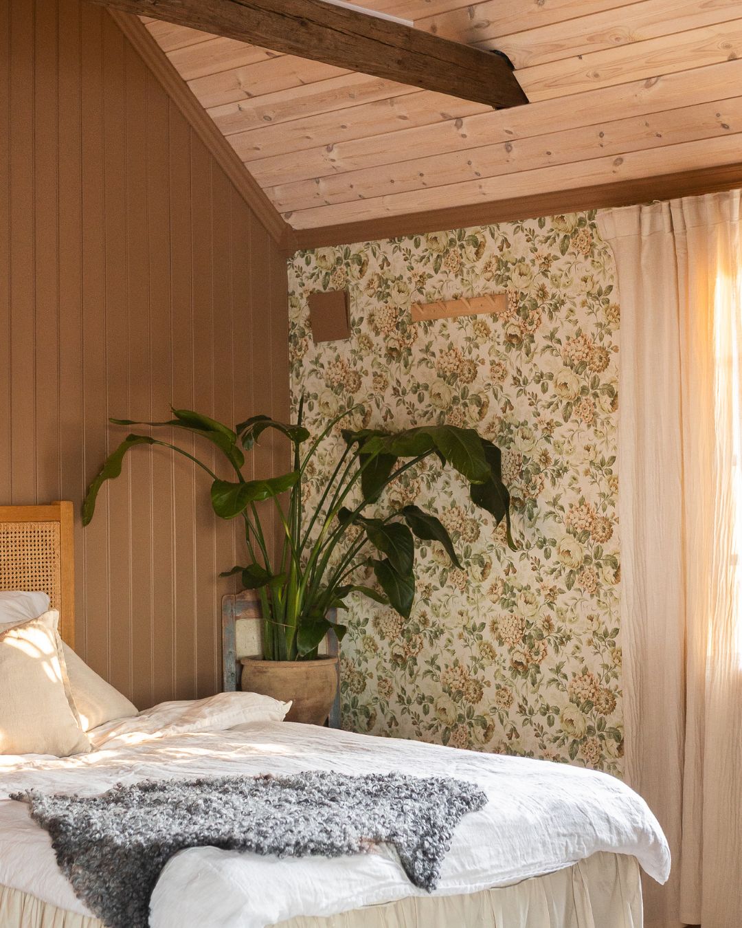 Sovrum med ockragul pärlspont på väggen och blommönstrad tapet bakom sängen. Rummet har vitbäddad säng, en stor grön krukväxt och ett ljust trätak med synlig takbjälke.