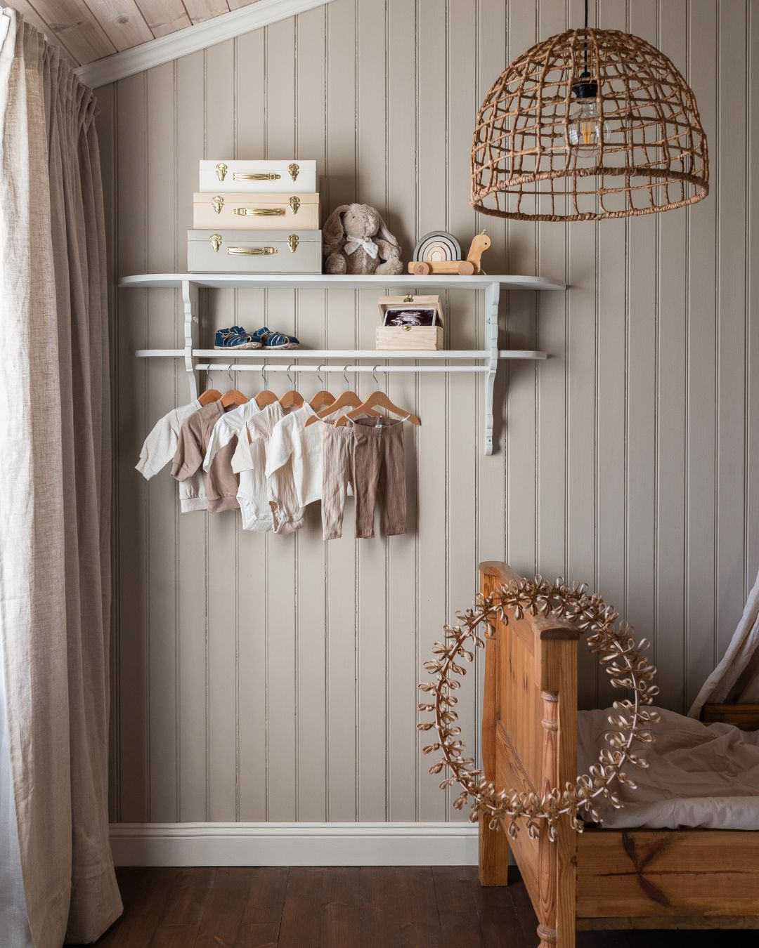 Barnrum med beige pärlspont på väggen och en vit vägghylla med barnkläder på galgar. På hyllan står förvaringslådor, en mjukiskanin och leksaker, och bredvid syns en träsäng samt en taklampa i flätad naturfiber.