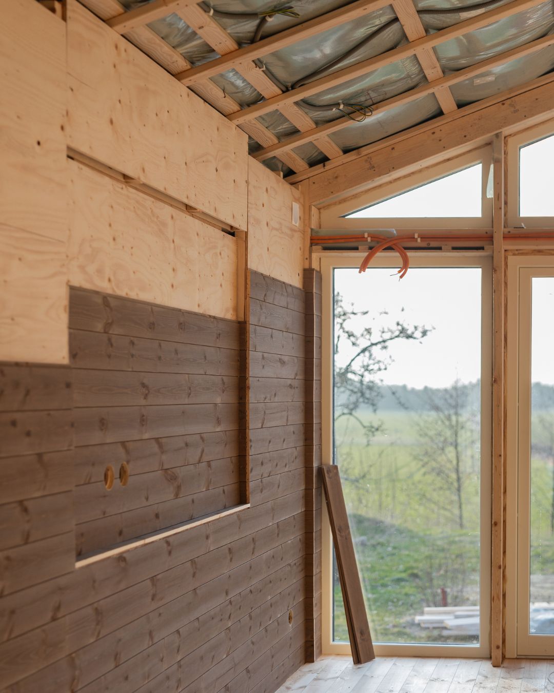 Rum under renovering med mörkbetsad träpanel monterad på delar av väggarna och bakom sitter plywood.. Taket är öppet med synlig isolering och reglar, och stora fönsterpartier släpper in dagsljus och utsikt över landskapet utanför.