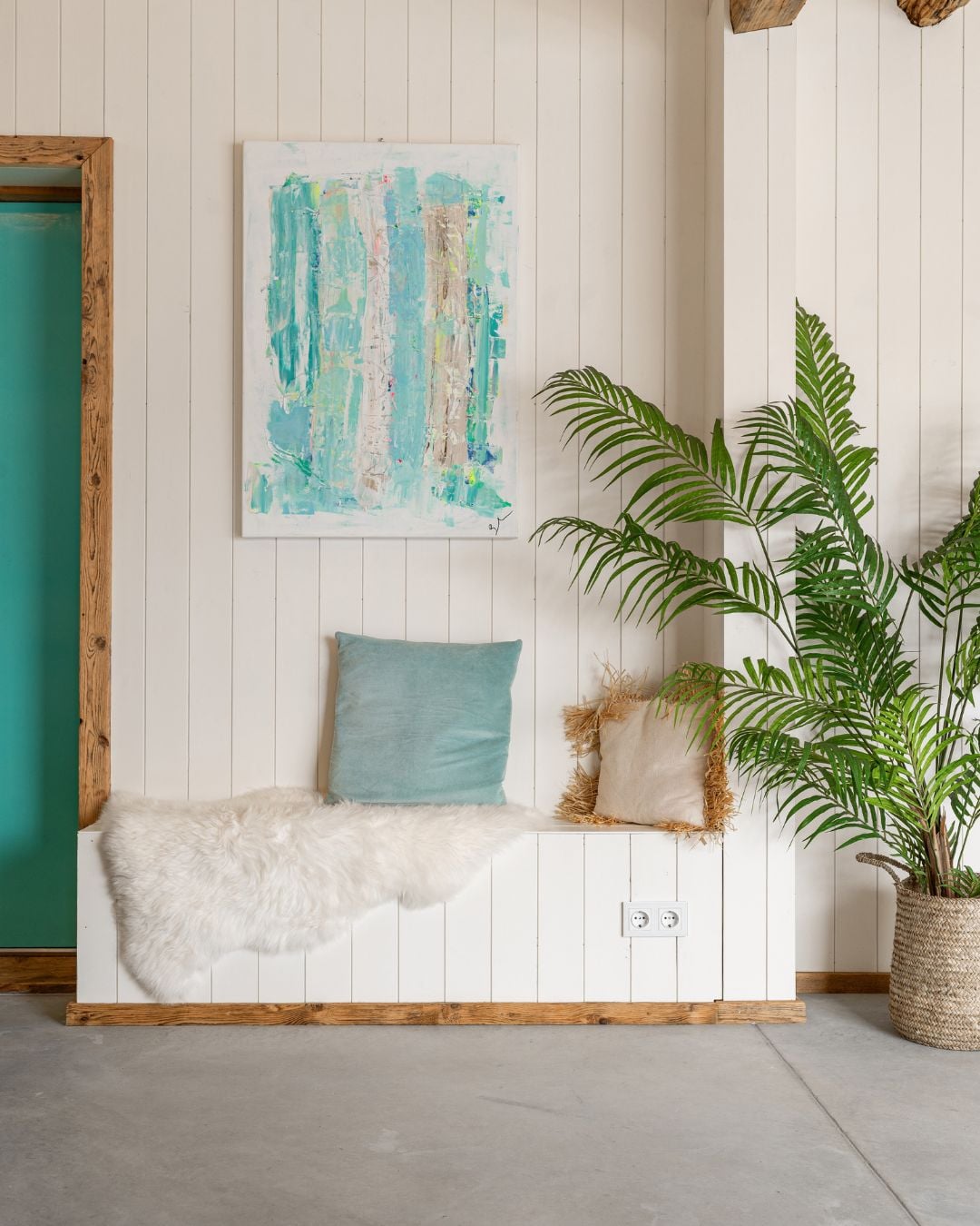 Vit sittbänk med skinnsfäll och kuddar längs en vägg med vita träpanelen Trend Slät och färgglad tavla och en stor växt