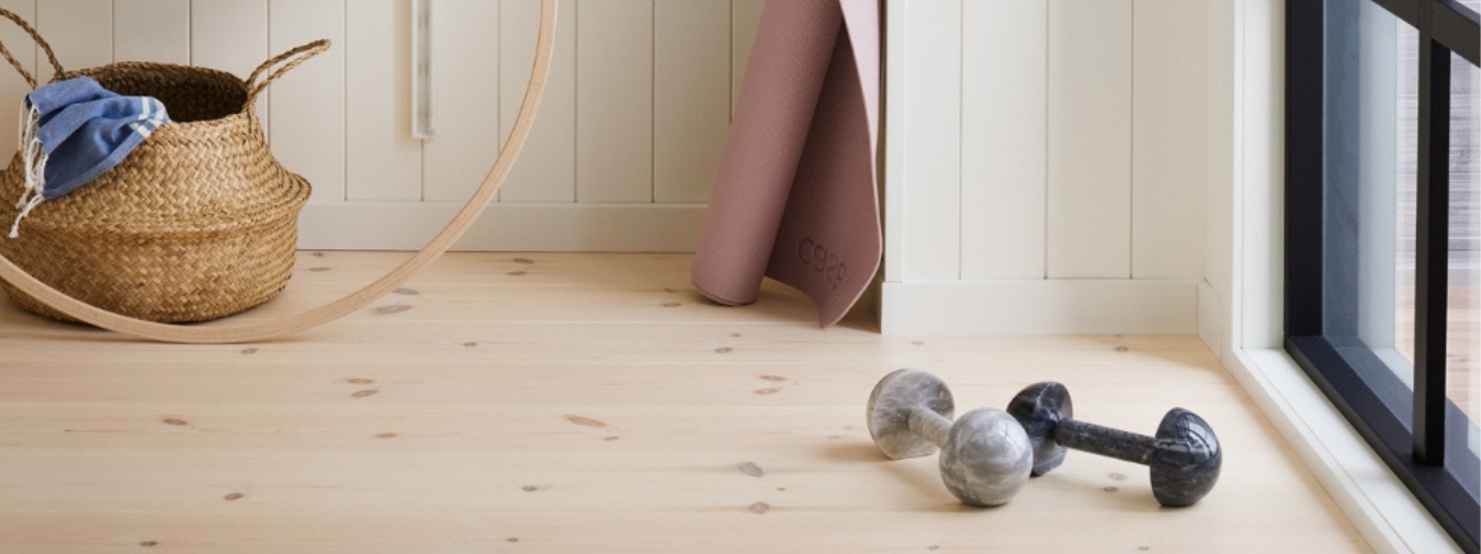 Ett hemmagym med hantlar och pilatesmatta. Ljust trägolv och vit träpanel