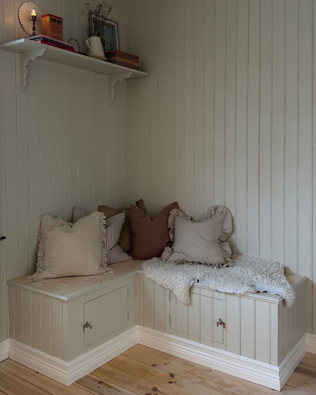 Färdigbyggd kökssoffa placerad i ett hörn av rummet. Soffan är klädd med stående panel i samma kulör som väggarnas pärlspont och har sitsar i ljust trä. På sitsarna ligger flera kuddar i ljusa och dämpade färger. Ovanför soffan sitter en vit vägghylla med böcker, en korg och andra inredningsdetaljer.