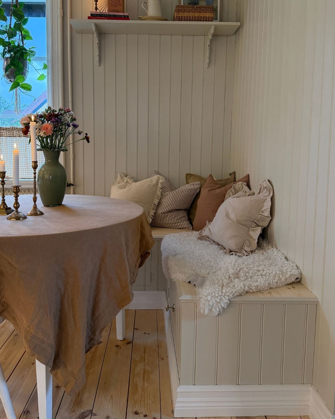 Kökssoffa med sitsar i ljust trä och förvaring bakom luckor i fronten. Soffan står intill ett matbord med duk, ljusstakar och en vas med blommor. På väggen ovanför soffan sitter en vit hylla med böcker och korgar, och vid fönstret hänger en grön växt. Rummet har väggar klädda med pärlspont och ett ljust furugolv.