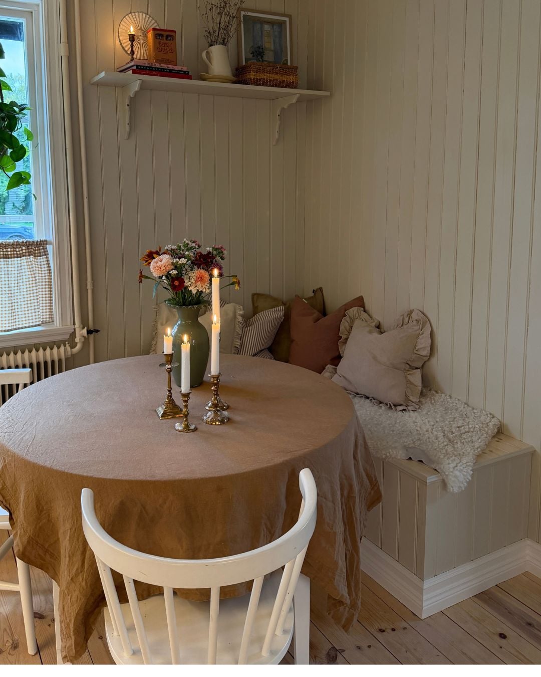 Ett runt matbord med duk står framför en platsbyggd kökssoffa i ett hörn av rummet. Soffan är klädd med stående panel, har sitsar i ljust trä och är dekorerad med kuddar i olika färger samt ett vitt fårskinn. På bordet står en vas med blommor och tre tända ljus. Intill soffan syns ett fönster med en grön växt, och ovanför soffan sitter en vit hylla med böcker, en korg och andra inredningsdetaljer.