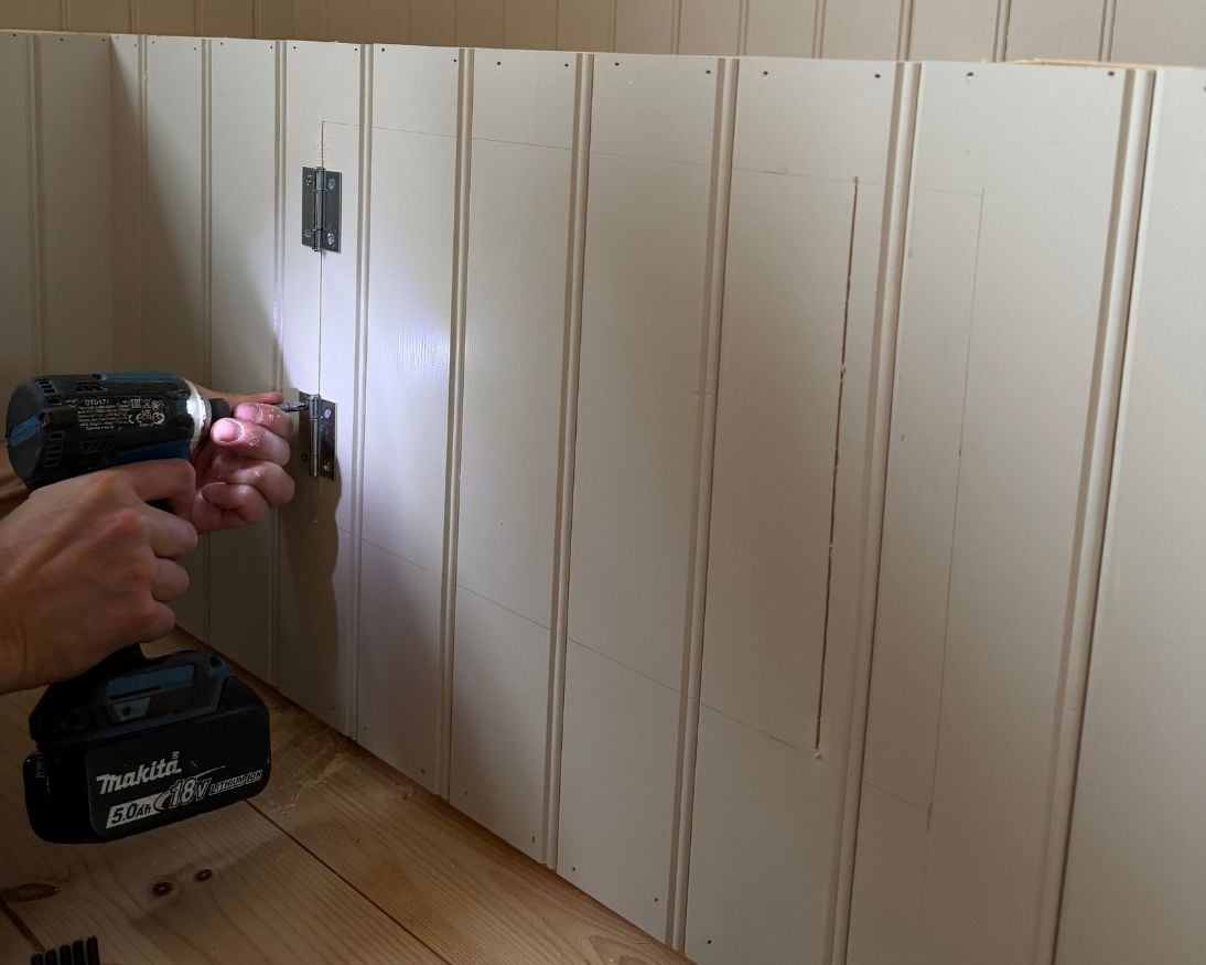 En person skruvar fast gångjärn på en utsågad lucka i panelen på framsidan av en bänkstomme. Luckans konturer är markerade i panelen och den övre delen av stommen är fortfarande öppen.