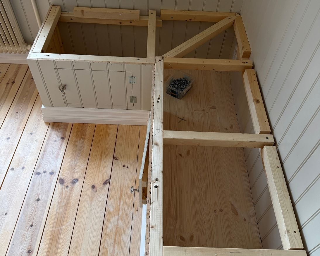 Bänkstomme sedd ovanifrån med öppna förvaringsfack under sittdelen. Framsidan är klädd med panel och försedd med luckor, medan extra tvärgående reglar har monterats i stommen för att ge stöd åt den kommande sittdelen. På golvet intill ligger en ask med skruvar.