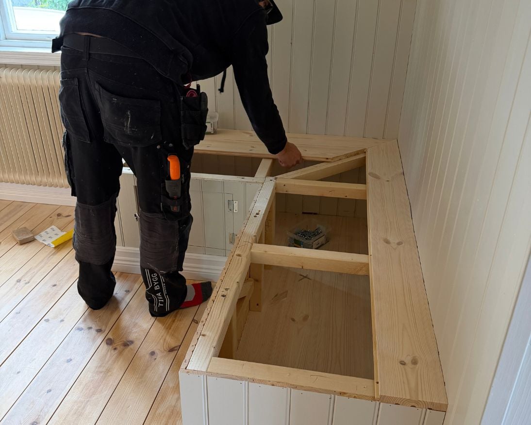 En person monterar golvbrädor som sittyta på en bänkstomme i trä. Delar av sittytan är redan på plats, medan ett öppet förvaringsfack fortfarande syns i mitten av konstruktionen. Bänken är klädd med panel och står mot väggar med pärlspont i ett rum med ljust furugolv.