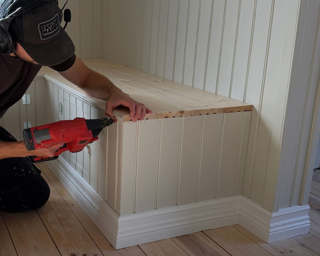 En person fäster en smal dekorlist längs den övre kanten av en bänk klädd med stående panel och sittlock av golvbrädor. 