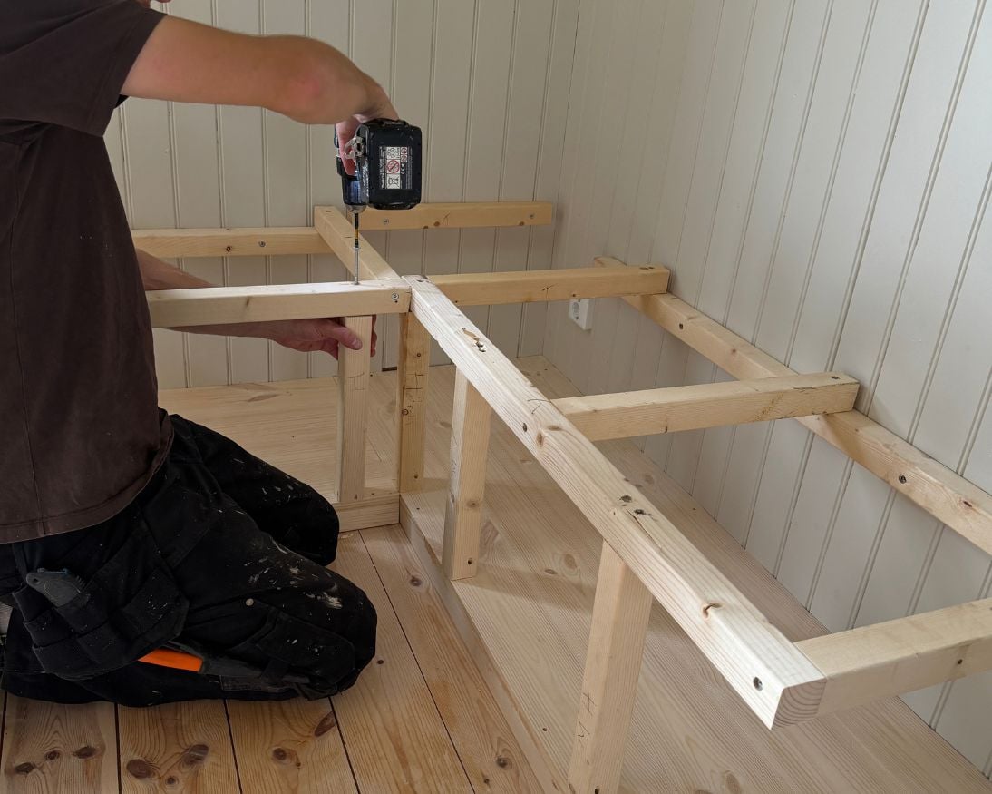 En person skruvar fast vertikala stödreglar på framsidan av en trästomme som är monterad mot två väggar med ljusgrå pärlspont. Den nedre delen av konstruktionen är klädd med golvbrädor.