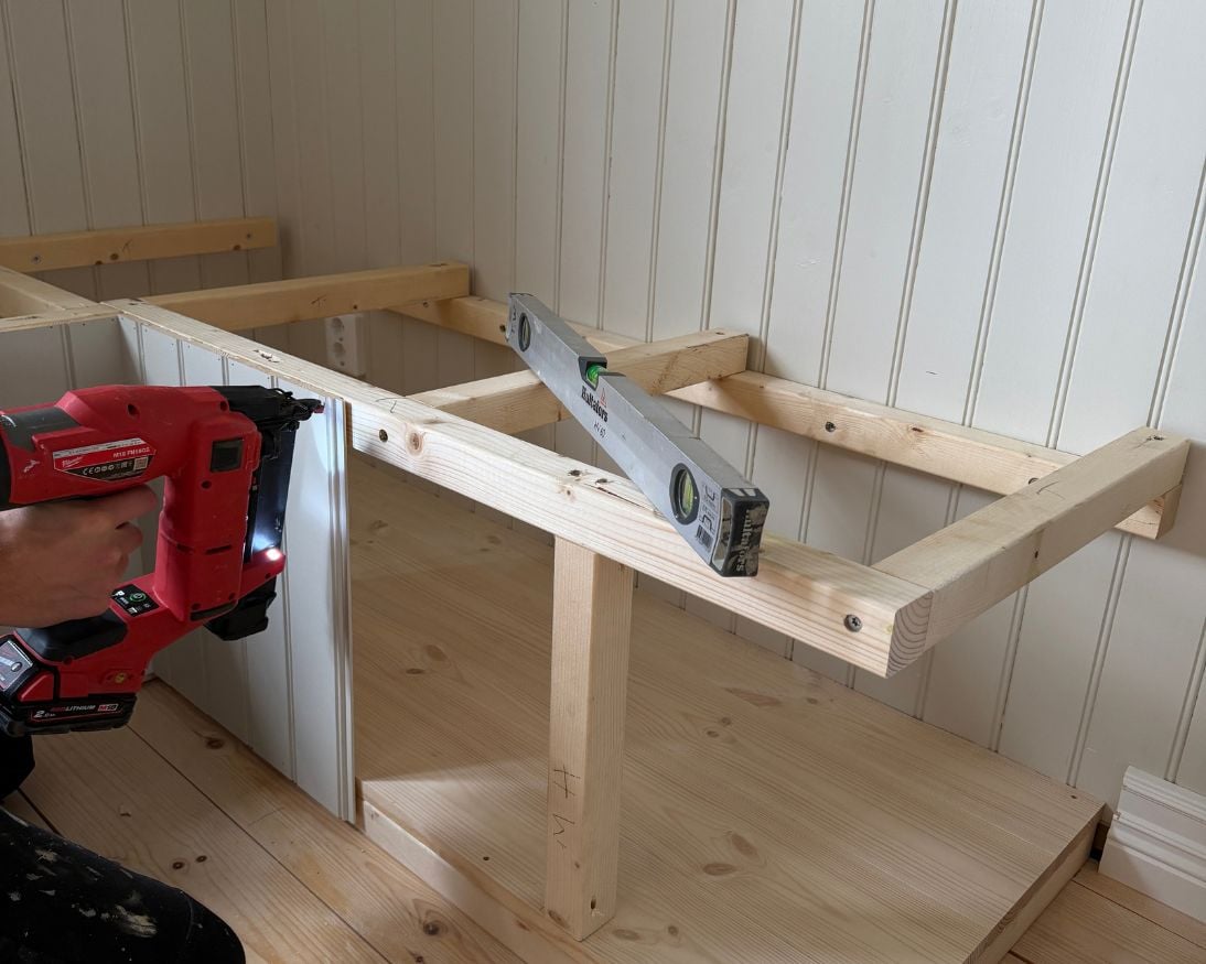 En person monterar stående panelbrädor på framsidan av en trästomme med hjälp av en spikpistol. Ett vattenpass ligger ovanpå stommen, och den nedre delen är klädd med träpanel.