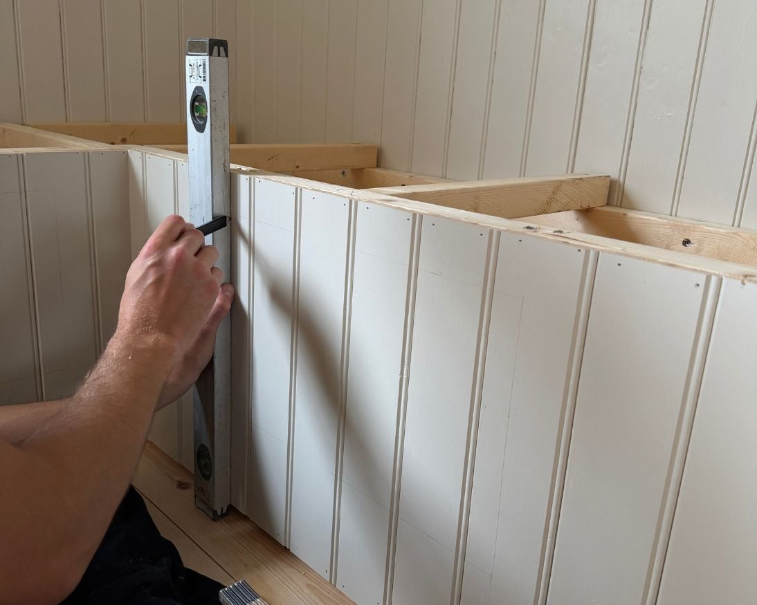 En person håller ett vattenpass mot framsidan av en bänkstomme klädd med vitmålad stående panel. På ovansidan syns den öppna trästommen som ännu inte har fått sittdel eller lock monterat.