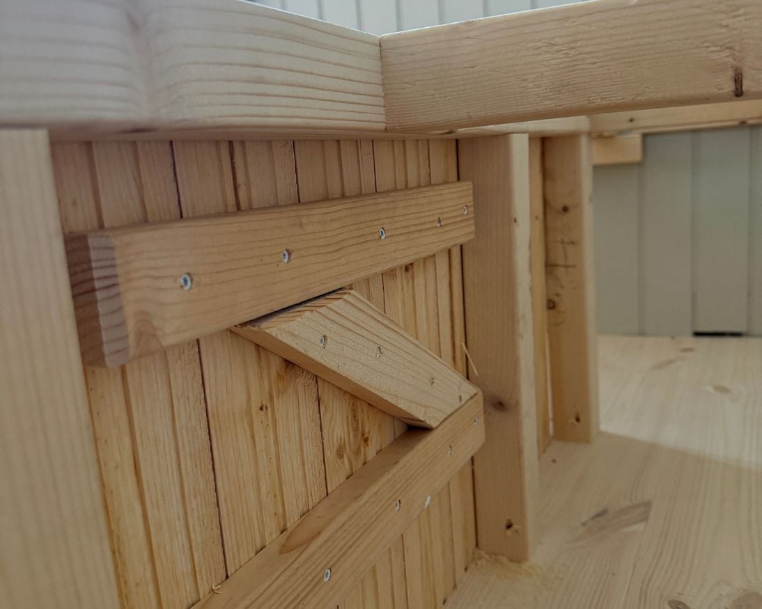 Insidan av en öppnad lucka i en bänkstomme. Luckan är byggd av träpanel och förstärkt med tvärgående och diagonala träreglar på baksidan. Ovanför syns den öppna trästommen som ska bära sittdelen.