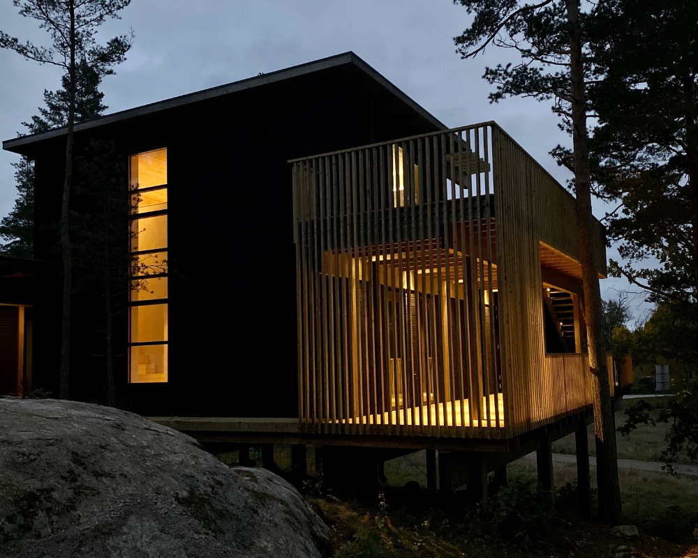 Ett hus med svart limträpanel i skogen, mörkt och lmapr i huset lyser