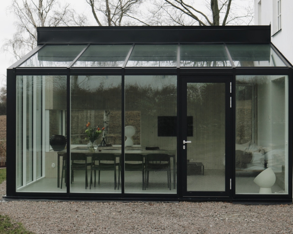 Ett inglasat hus med svart ram, i huset finns bord och stolar och en tv, samt övriga inredningsdetaljer