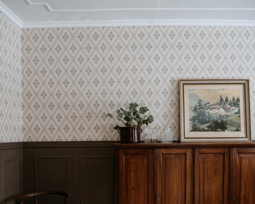 Ett rum med mörk bröstpanel, en brun byrå, mönstrad tapet, tavla och blomma