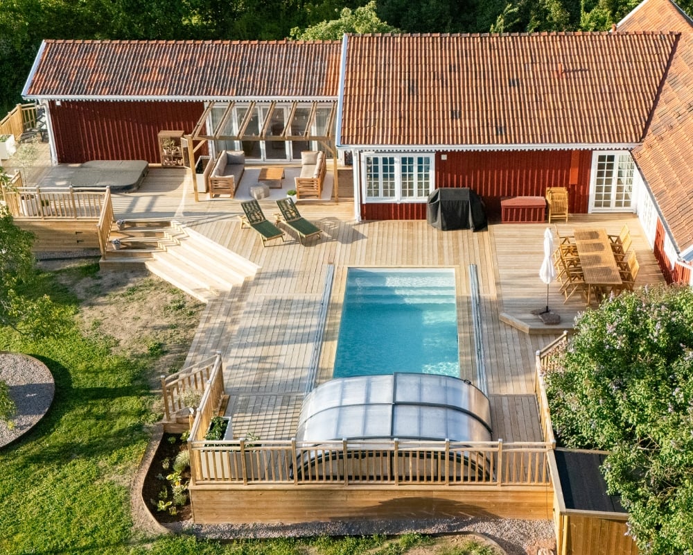 Ett rött hus och en stor altan i trä, pool, jacuzzi och utomhusmöbler, samt en tomt med gräs och träd