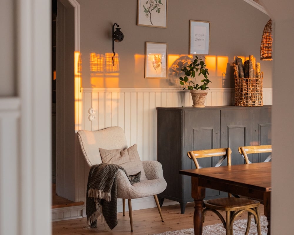 Matsal med vit bröstpanel i pärlspont. Ett brunt matbord och stolar i trä. En fåtölj teddypläd  och ett grått skåp, samt dekorationer