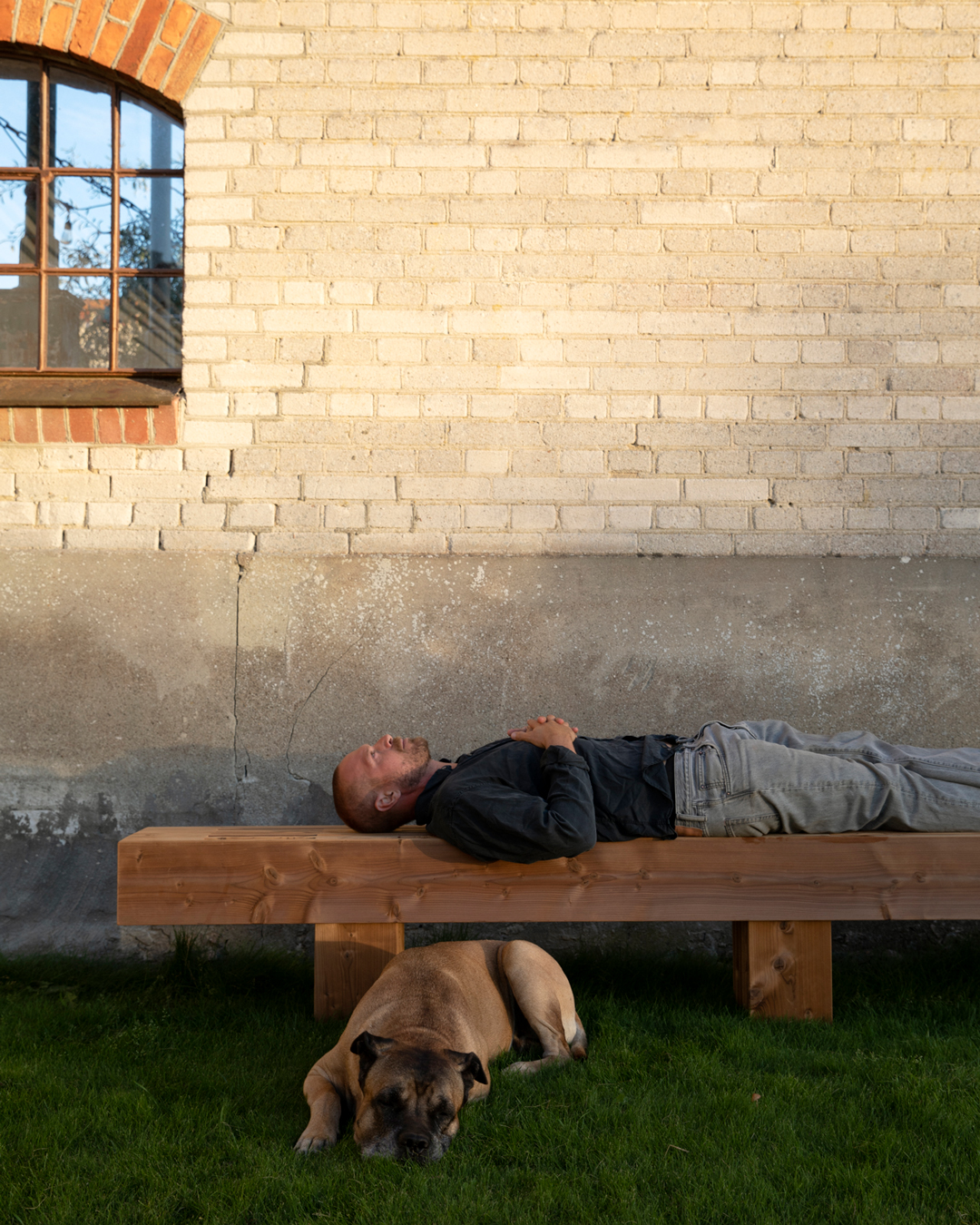 Gustav Ovland ligger på en platsbyggd träbänk i TermoGran framför fabrikens fasad, med en av hans hundar vilandes på gräset bredvid.